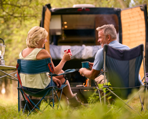 Senior Couple Camping In Countryside With RV Drinking Coffee By Outdoor Fire